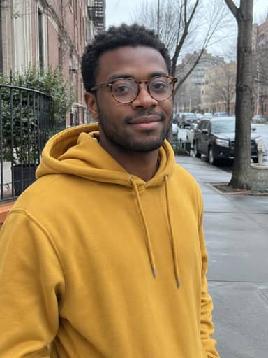 Uploaded photo: A street photo in a mustard hoodie. Same round glasses and stubble, with stars in his eyes.